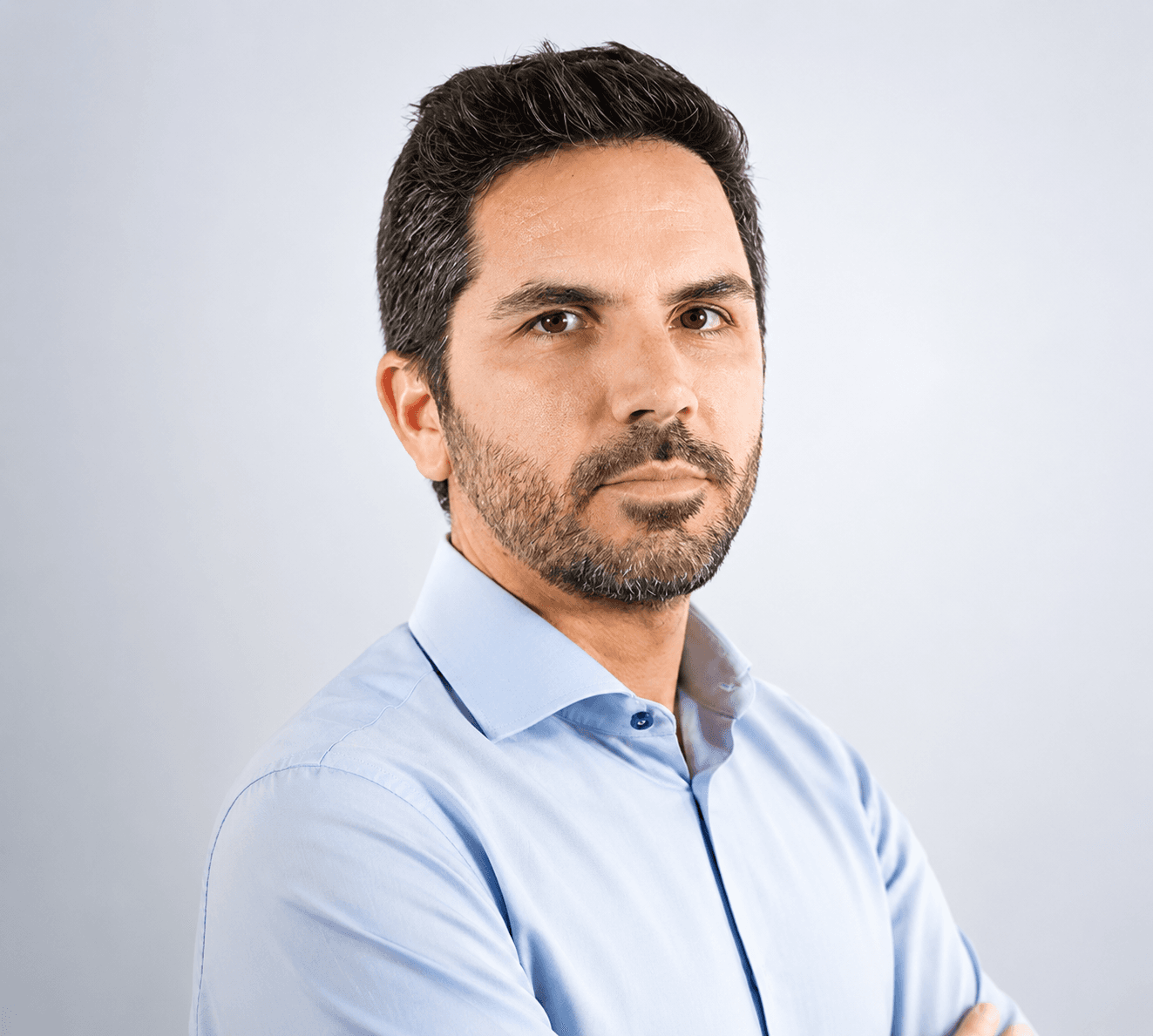 Uomo con barba e capelli scuri indossa una camicia azzurra su sfondo grigio chiaro.