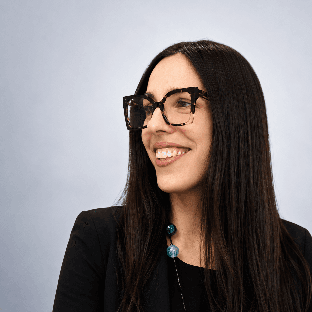 Donna sorridente con lunghi capelli scuri, occhiali tartarugati e giacca nera, guarda di lato.